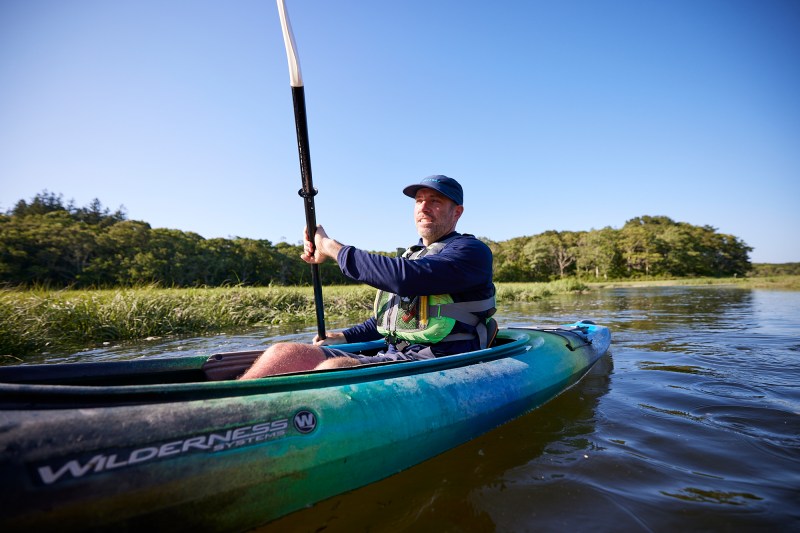 Man kayaking on a calm lake surrounded by green trees under a clear blue sky.