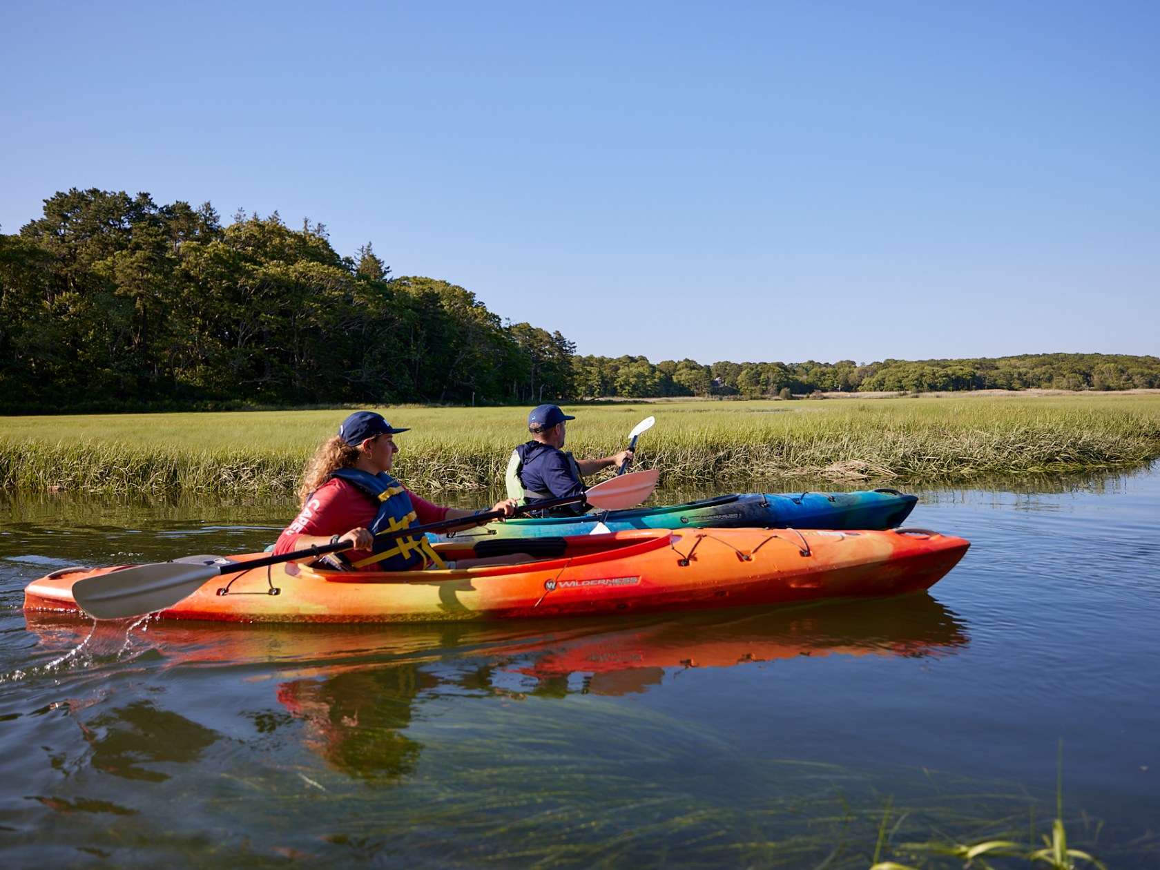 Two people kayaking on a calm river near a grassy shore under a clear blue sky.