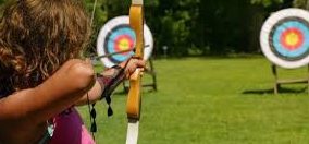 Person aiming a bow at a target on a grassy field.